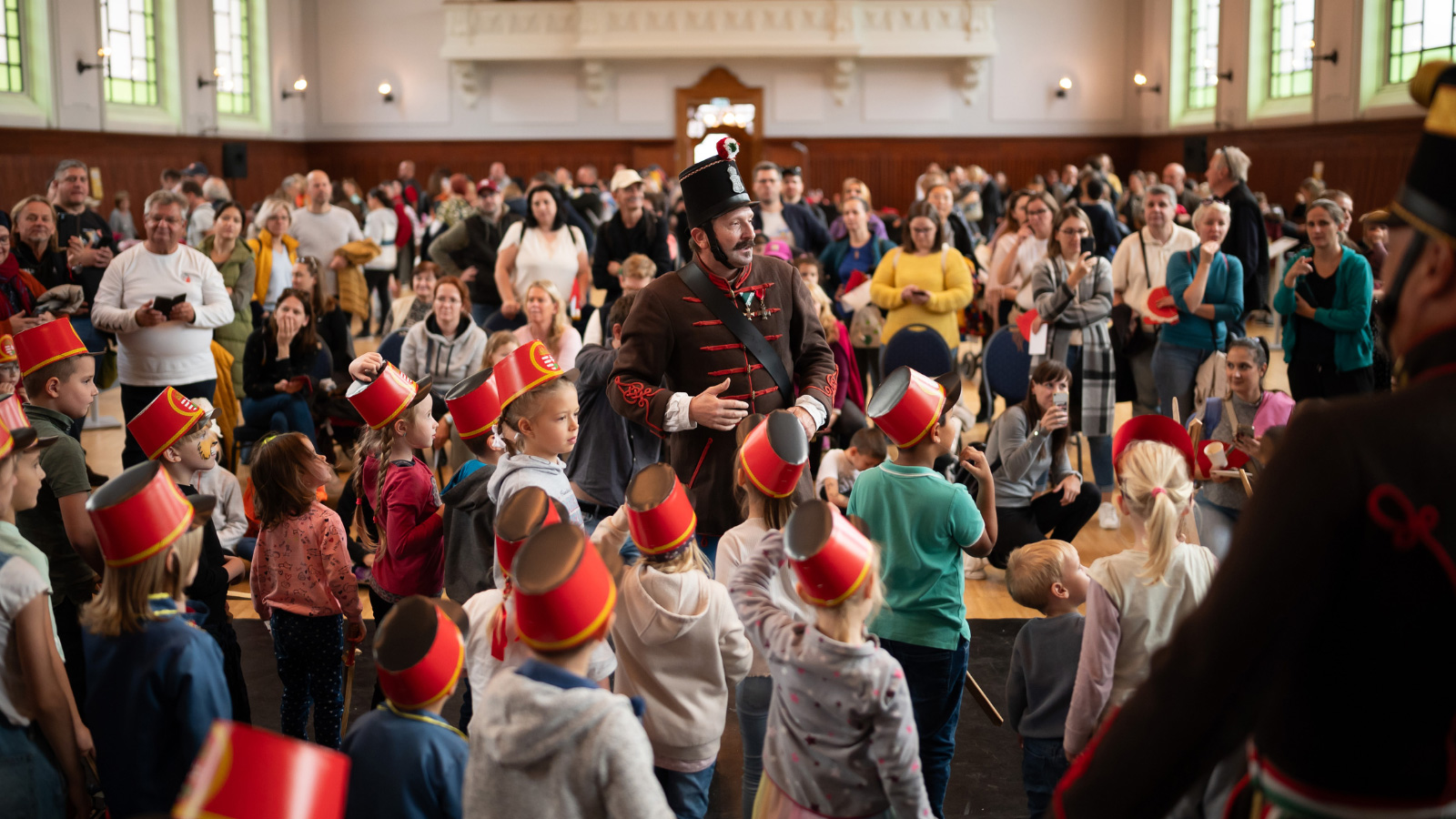 Férfi huszár ruhában áll, körülötte érdeklődő gyerekek és szülők egy csarnokban