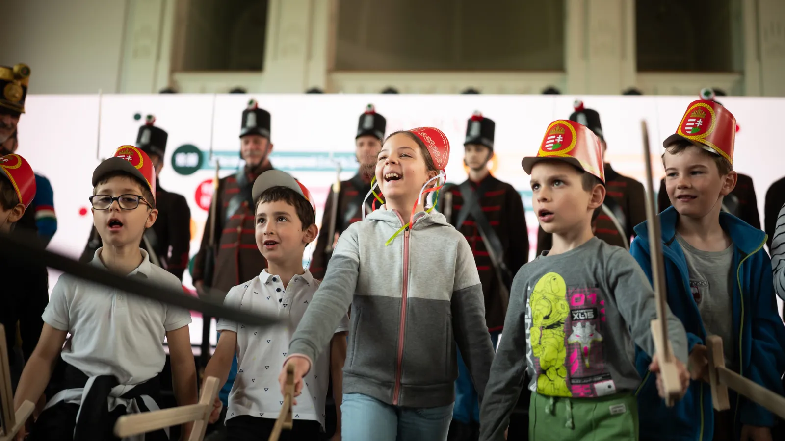 Gyereknapi programon gyerekek huszárcsákóban, fakarddal állnak magyar huszárok előtt